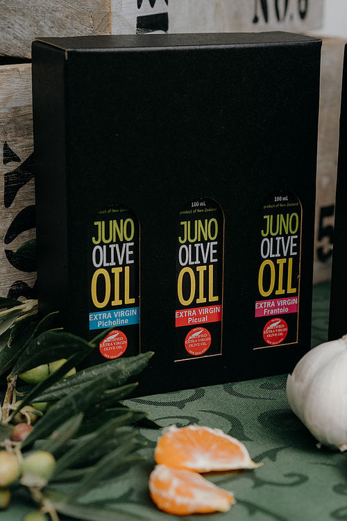 3 x 100 mL bottles of olive oil in a black gift box surrounded by fruit and olive branches.
